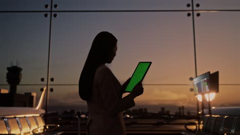 vista trasera de una mujer de negocios asiática con una maleta rodante en la sala de embarque del aeropuerto, usando una tableta de pantalla verde mientras espera el vuelo, el avión despega fuera de la ventana
