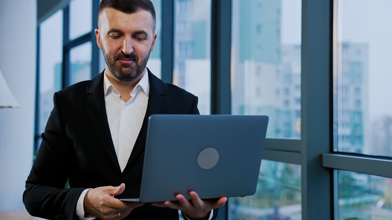 Smiling good-looking bearded businessman in suit stands near the window holding laptop. Man talks showing something with his hands.