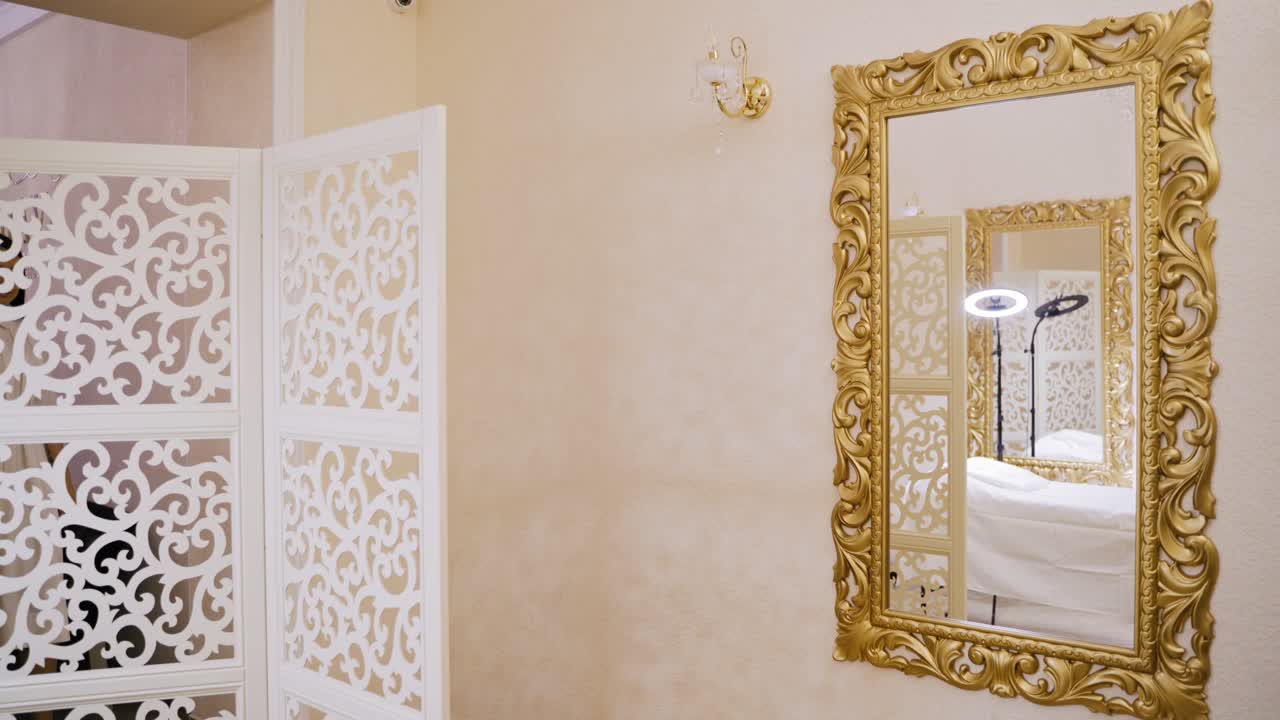 A woman in a white dress come in the room and sits on a couch opposite the mirror, waiting for the procedure to start at the cosmetology center.