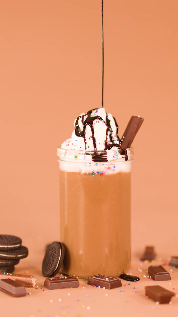 Sciroppo al cioccolato che cola su un milkshake con panna montata, biscotti, codette di zucchero e cioccolato, su sfondo marrone