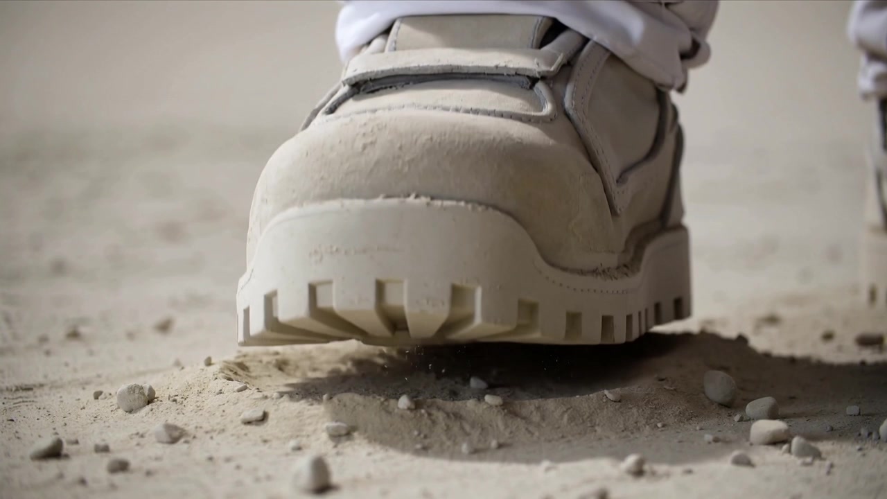 Menggeser berat badan, seorang pekerja mengangkat sepatu bot krem dari lantai berdebu dengan manset putih, menggoyangkan kerikil.