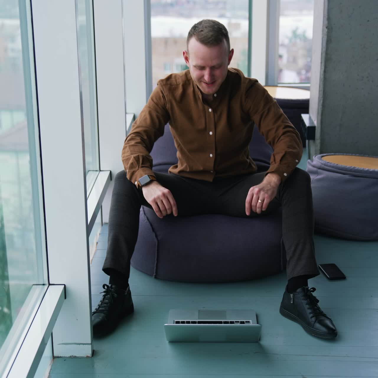 Young man with a laptop near the window. Handsome guy in shirt is sitting on a comfortable ottoman and looking on his laptop waiting for something