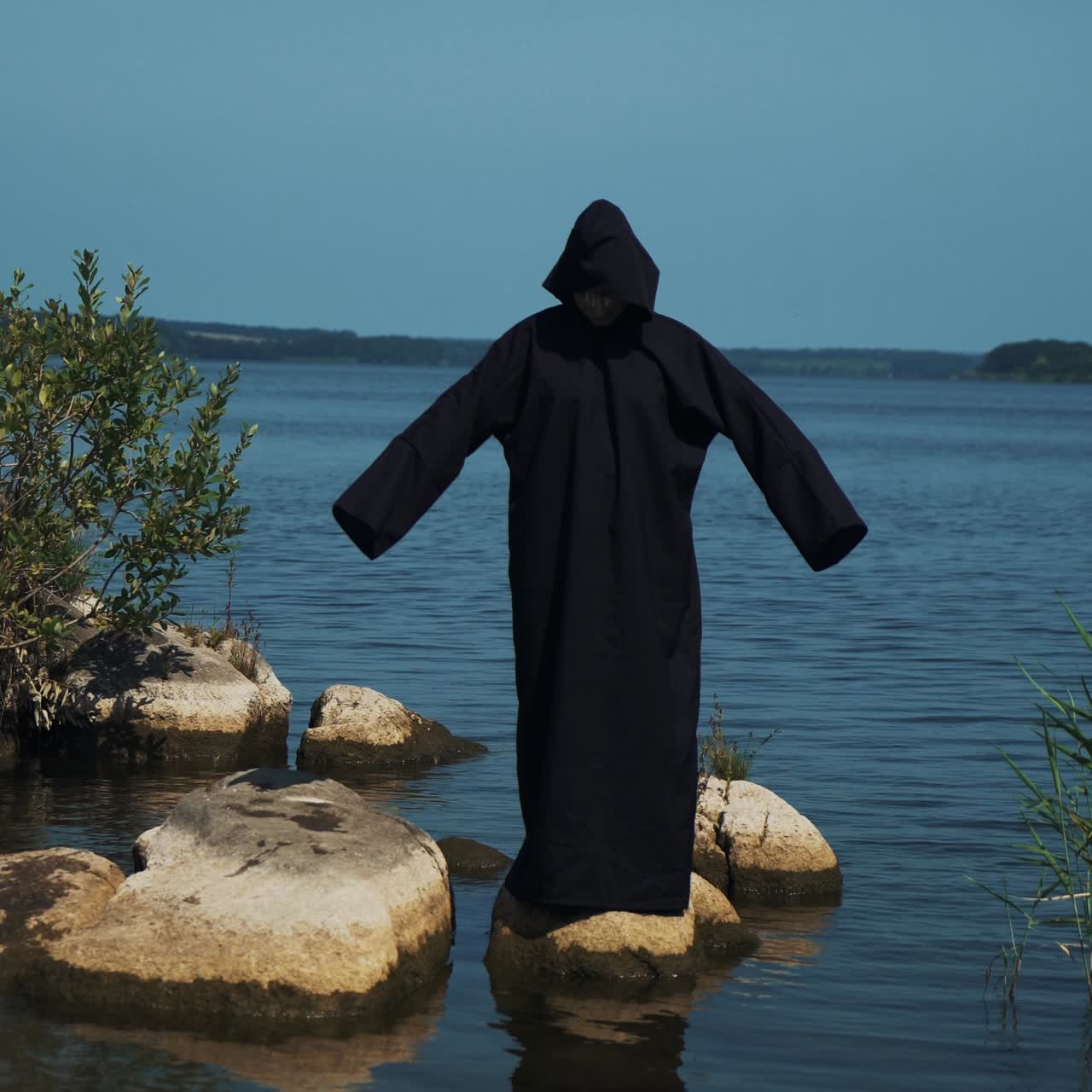 Woman devil in black cape and hood on stones near the river. Halloween costume.