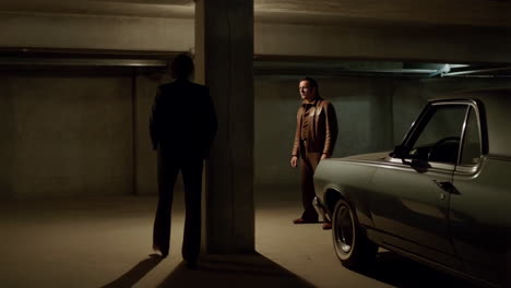 two men in a dark parking garage with a vintage car