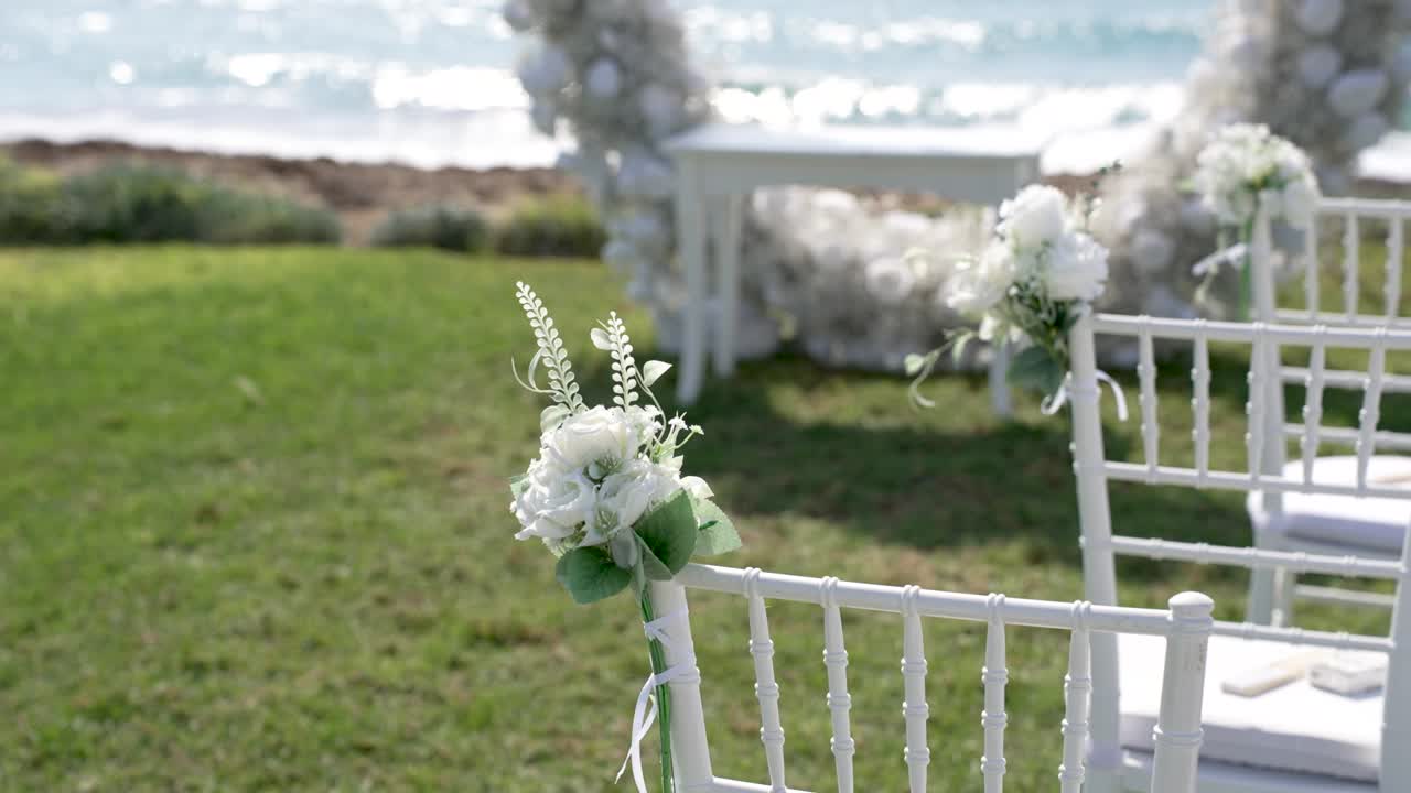 Delle sedie sono disposte su un prato verde vicino alla spiaggia. Fiori bianchi decorano le sedie mentre gli invitati si preparano per una cerimonia nuziale in riva al mare.