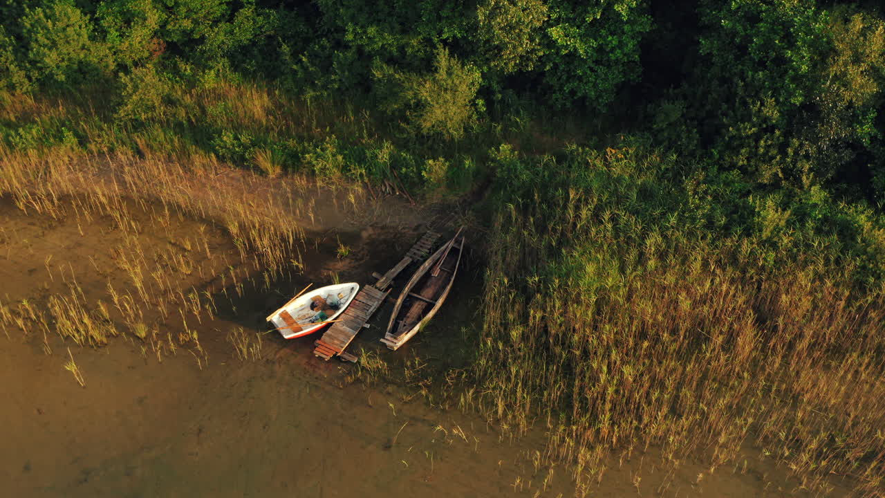 Two old wooden row boats rest by a small, damaged pier on a tranquil lake shore surrounded by reeds and lush green trees, high-angle drone shot.fi