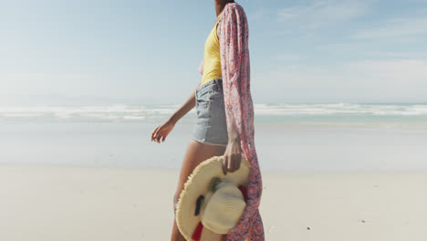 feliz mujer afroamericana caminando con sombrero en una playa soleada
