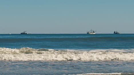 Long-shot-of-tug-boats-and-shrimping-boat-on-the-ocean-in-the-background-with-waves-crashing-in-the-foreground