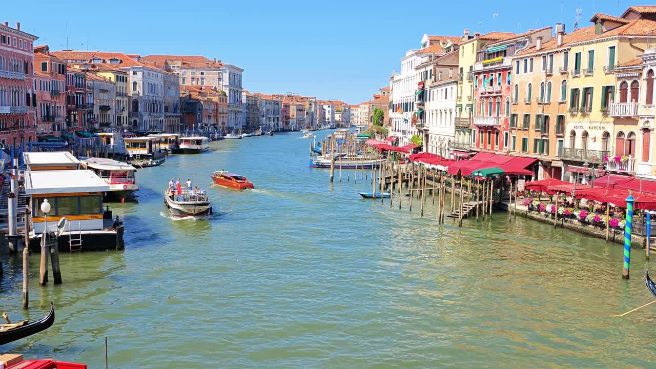 Canal with boats and gondolas passing by colorful historic buildings under a clear sky, tourists enjoying the waterfront view