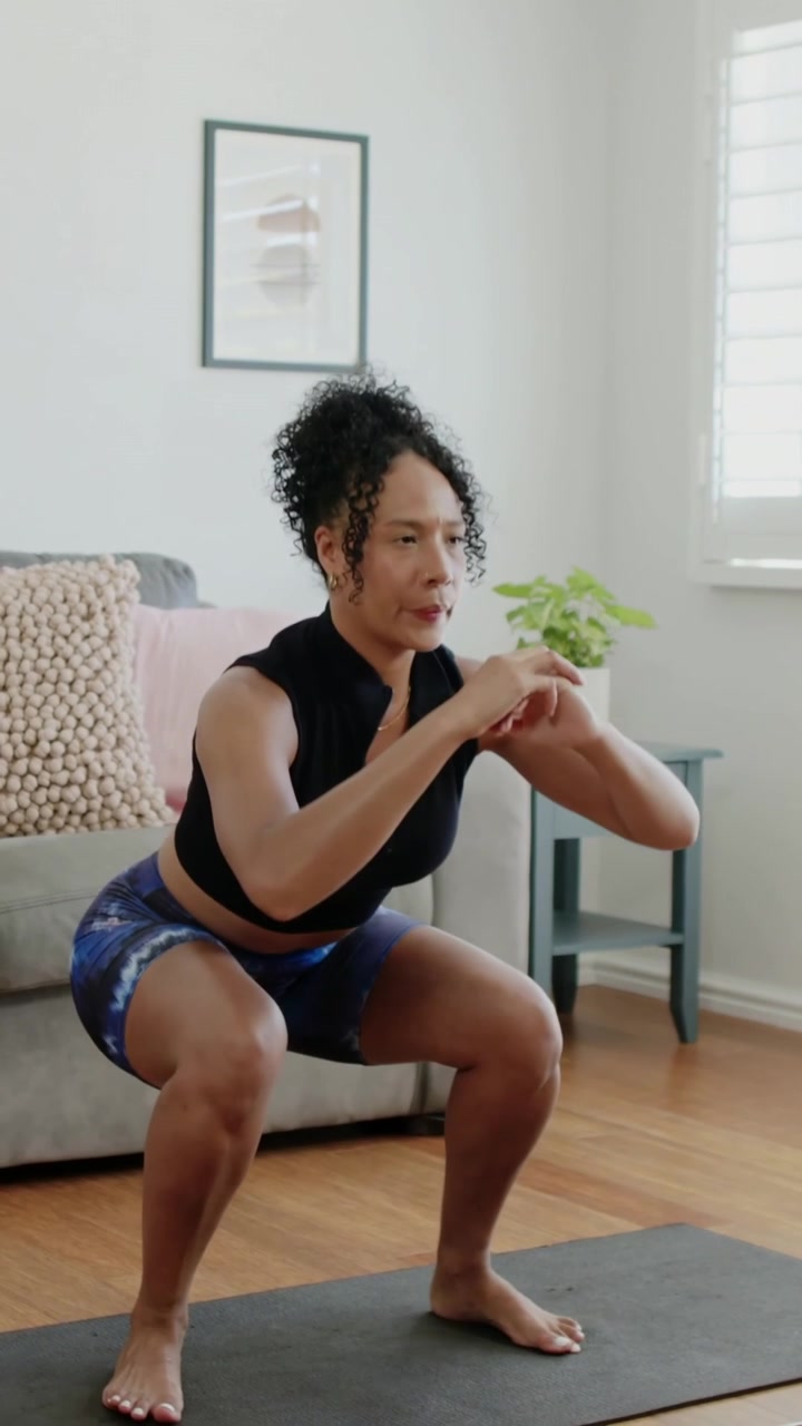 Video verticale: All'inizio dell'allenamento, una donna esegue ripetizioni di squat-to-stand a casa su un tappetino, indossando un top nero.