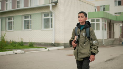 boy walks confidently after class holding book and tablet under arm with backpack on shoulder approaching home showing determination and responsibility in daily academic routine