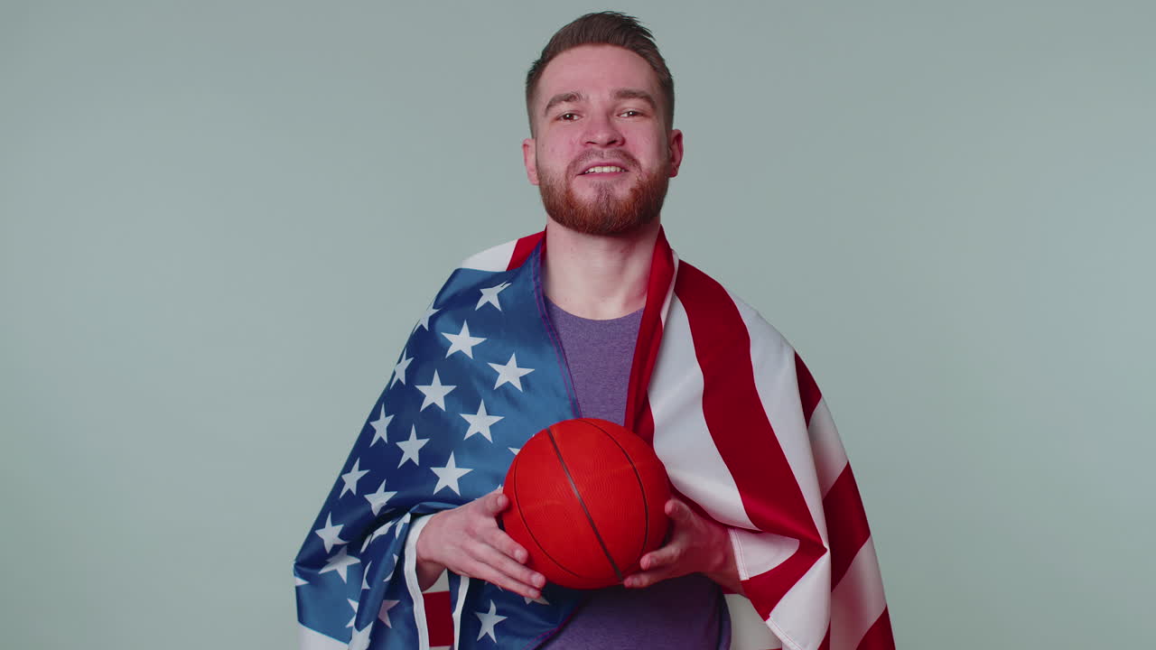 un joven barbudo fanático del baloncesto sosteniendo la bandera de los estados unidos haciendo el gesto del ganador, bailando solo