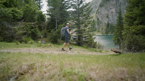 hiker with the hat, backpack and hiking poles walking towards the mountain lake, surrounded by conifers, mountain the back