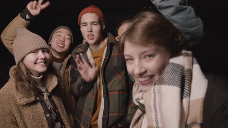 close up view of group of friends dressed in winter clothes waving at the camera at night