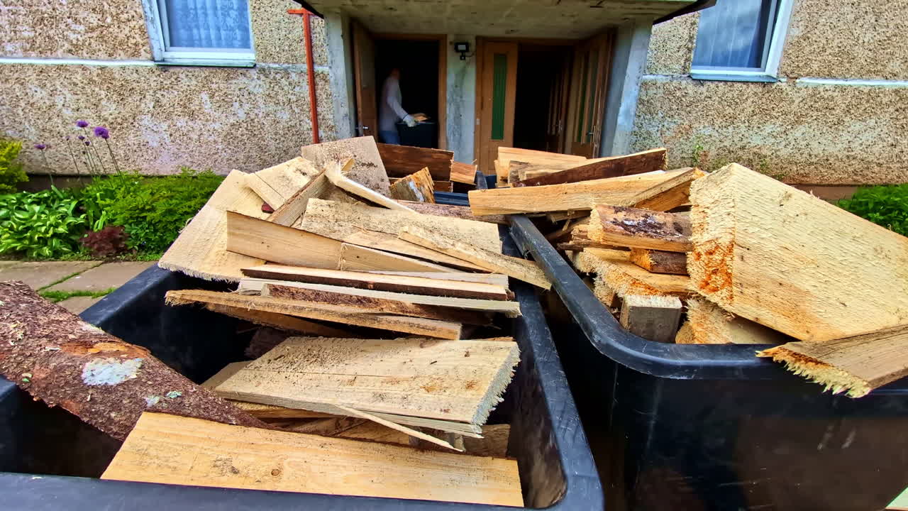 Firewood pieces in bins with person unloading from trailer at old house entrance