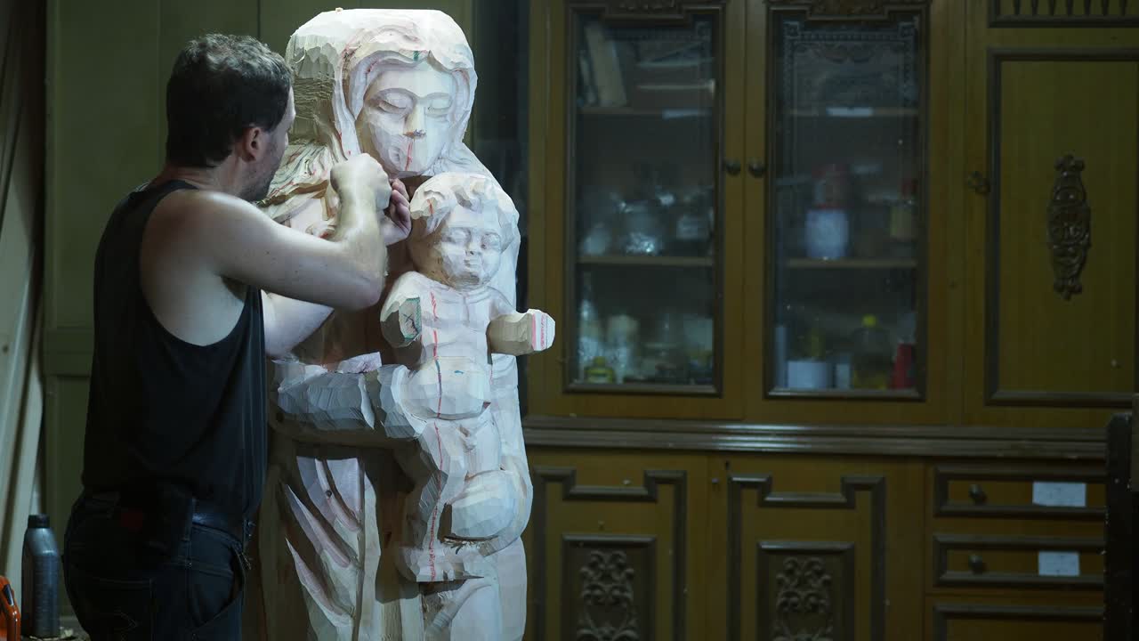 Italian adult artisan carefully chisels a large wooden sculpture of the Virgin Mary holding the Child Jesus, captured in slow motion inside a traditional studio with wooden cabinetry