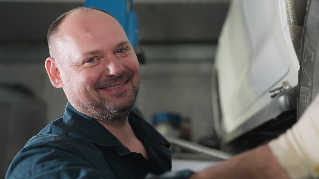 ingeniero en uniforme azul con guantes blancos sonriendo mientras alinea el neumático del coche para un rendimiento óptimo en el taller de reparación de automóviles, usando herramientas para la precisión