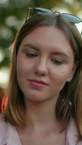 primer plano de una mujer joven mirando contemplativamente con una cálida sonrisa y gafas apoyadas en su cabeza, centrándose en algo delante de ella, el fondo presenta árboles y la luz filtrándose a través de ellos