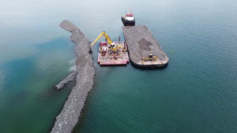excavadora en barcaza en agua construyendo rompeolas de rocas cerca de la costa