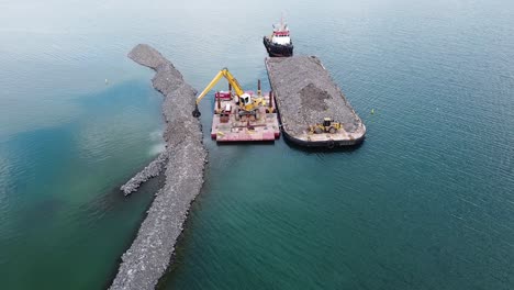 excavator on barge in water building breakwater from rocks near shoreline