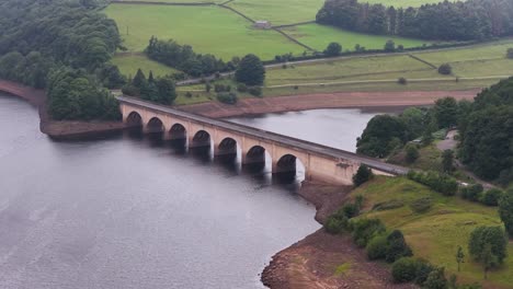 drone footage glides above a multi-arched stone bridge spanning a calm reservoir, surrounded by lush green fields and forest under soft daylight