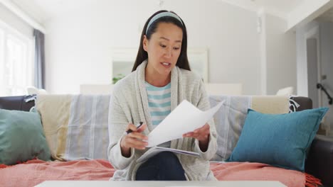 Asian-woman-working-at-home,-sitting-on-couch-making-video-call,-talking-and-reading-paperwork