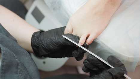 technician wearing black gloves carefully files toenails of client resting leg on cushioned surface during professional pedicure session, showcasing precision and hygiene in clean salon setting