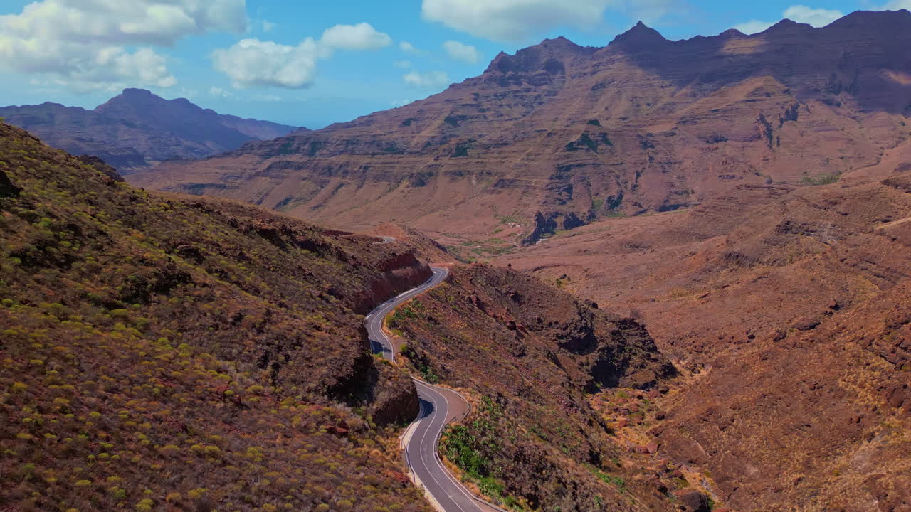 Ripresa con drone in avanzamento su una strada di montagna tortuosa a Gran Canaria, Isole Canarie, Spagna. Una strada asfaltata vuota si snoda attraverso un drammatico paesaggio vulcanico sotto un cielo blu con nuvole sparse