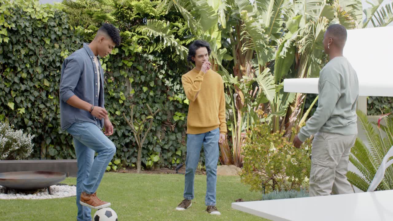 Diverse male friends rolling soccer ball in backyard pausing for chat then juggling ball practice