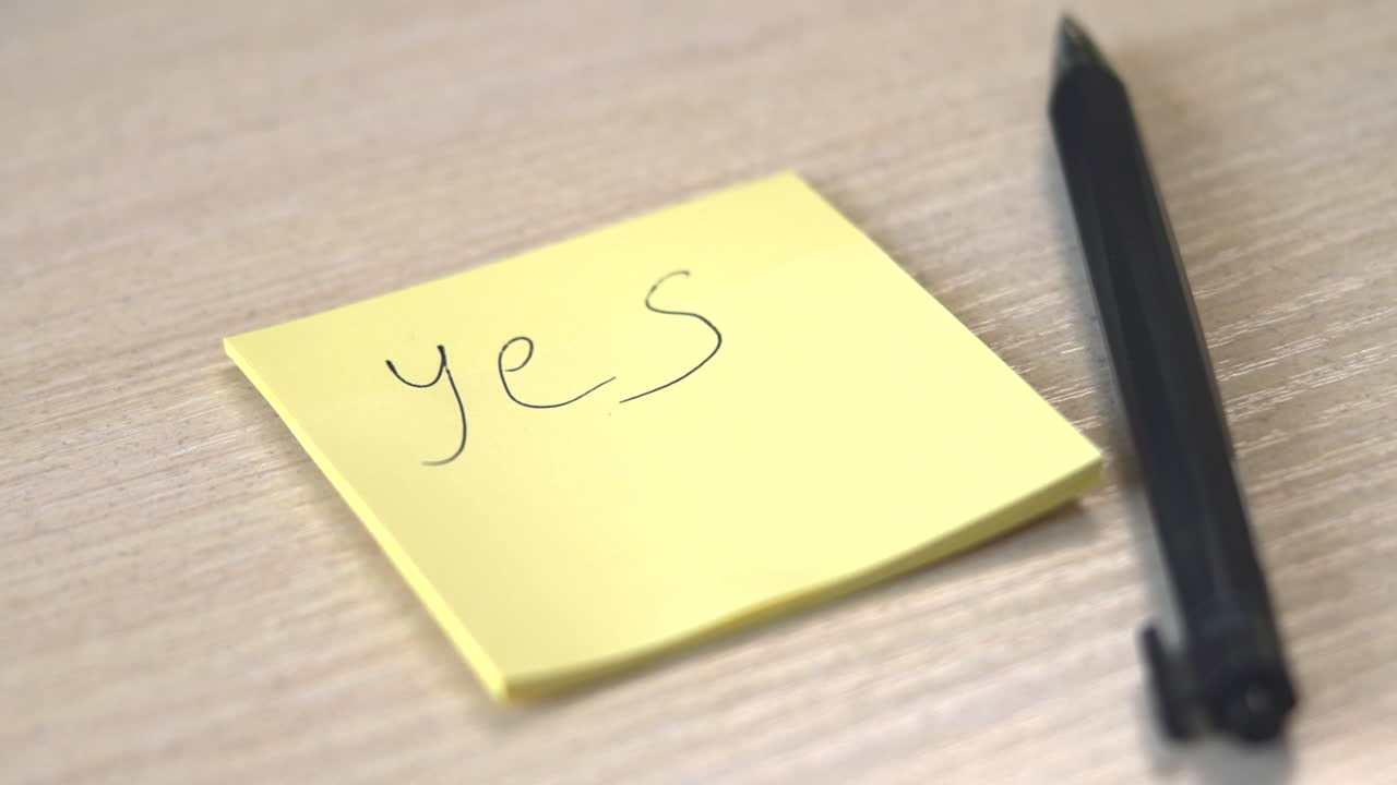 Post-it giallo con la scritta "Sì" su scrivania in legno accanto a penna nera, con profondità di campo ridotta e luce naturale soffusa