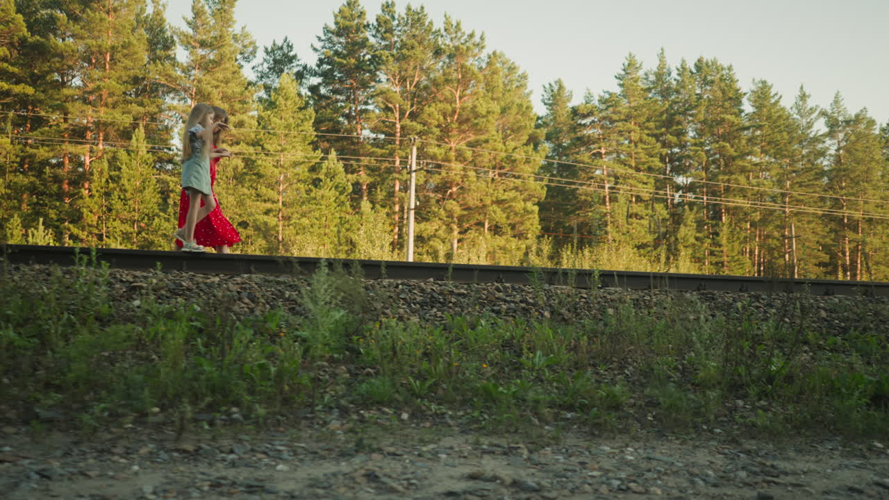 side view of woman in red dress walking beside young daughter on rail track through wooded area, background showing power lines and tall pine trees under clear sky during calm evening walk