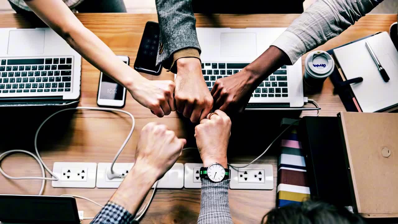 Top-down video shot of diverse hands fist-bumping over a wooden desk with laptops