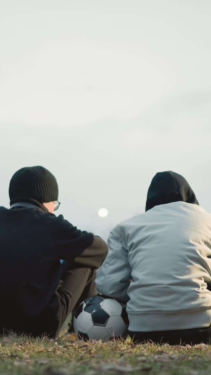 Two boys are seated on a grassy field with their backs to the camera, one in a gray jacket resting his left hand on a soccer ball, with a blur background