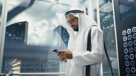 un hombre de negocios musulmán exitoso en el tradicional kandura blanco montando un ascensor de vidrio a la oficina en un centro de negocios moderno. hombre usando un teléfono inteligente. saudi, emirati, concepto de hombre de negocios árabe.