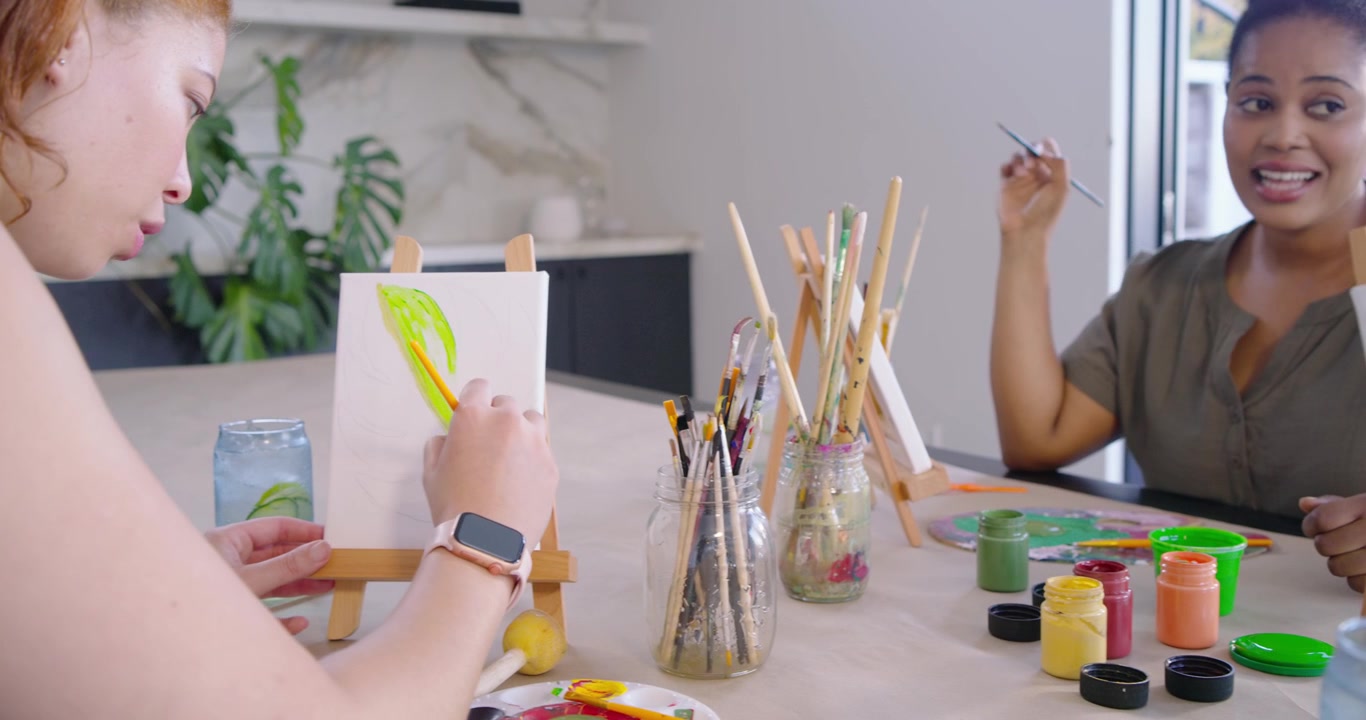 Mulheres diversas pintando à mesa, uma com blusa verde-oliva mergulhando a esponja na tinta verde e carimbando a tela.