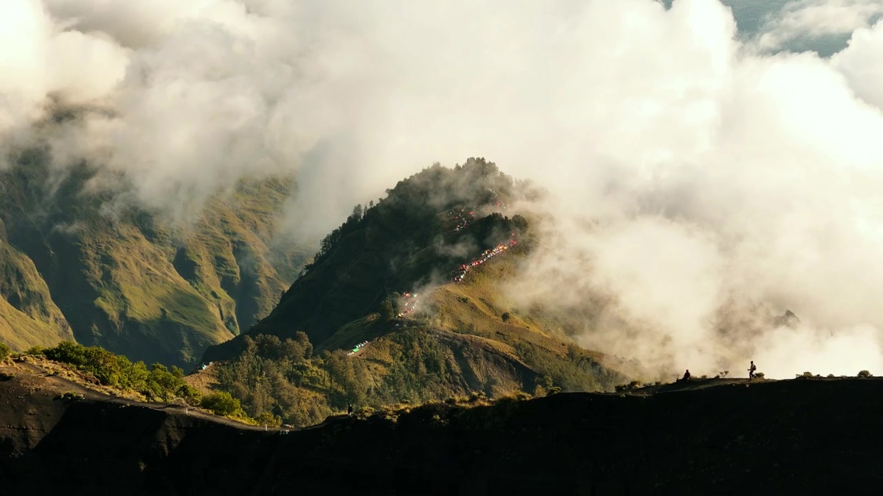Nuvole eteree si muovono sulle creste del Monte Rinjani sopra lussureggianti pendii vulcanici nella luce dorata del mattino, ripresa aerea in allontanamento.