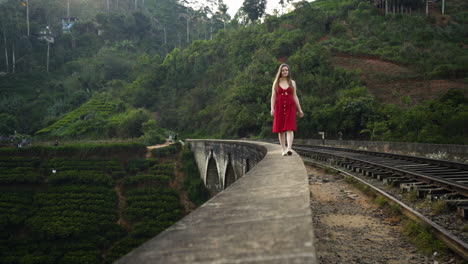 Nine-Arches-Bridge-Sri-Lanka,-A-young-tourist-girl-walks-along-the-rail-tracks-in-a-red-dress