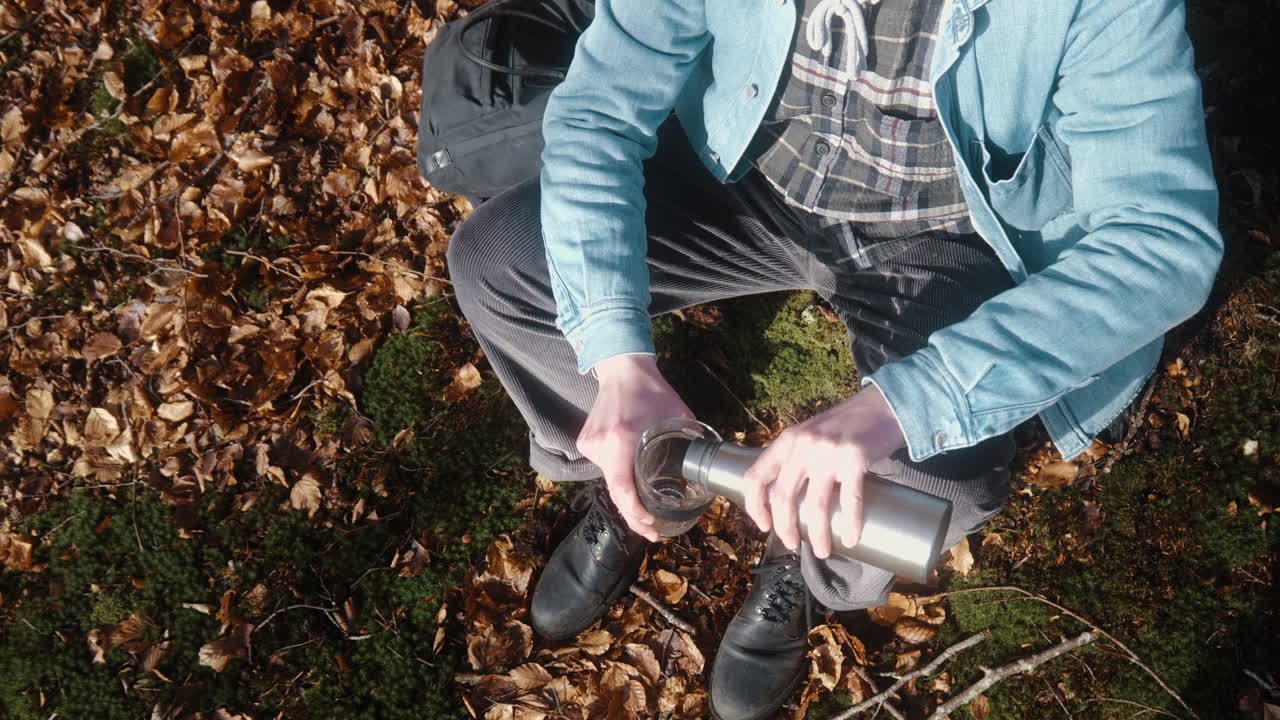 hombre sentado en el suelo del bosque, sirviendo café en su taza en un soleado día de otoño