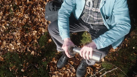 man-sitting-down-on-the-forest-floor,-pouring-coffee-in-his-cup-on-a-sunny-autumn-day