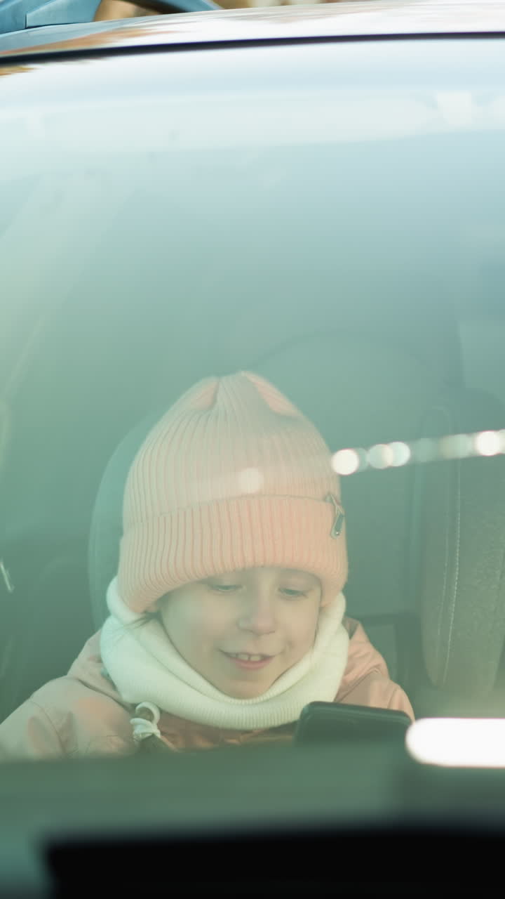 Inside a car, as a man in a green jacket sits in the driver s seat, while his daughter, dressed in a pink beanie and jacket, smiles at him while focused on a smartphone in her hands