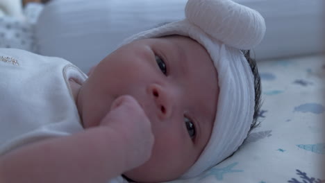 Baby-Girl-In-White-Headband-Lying-Awake-In-Bed-Putting-Hand-In-Her-Mouth