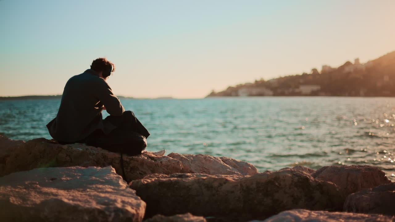 A man sitting alone on large rocks by the sea, looking toward the horizon under a clear sky