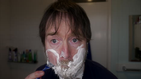 close up of caucasian man with deep blue eyes wet shaving his face with beard at home in bathroom