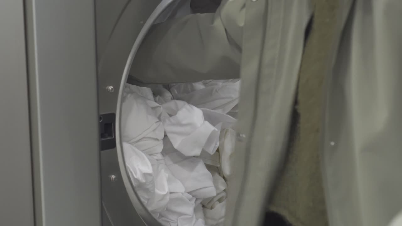 A detailed shot showing a person’s hands placing various pieces of dirty laundry into a modern commercial washing machine at a public laundromat, finally closing the glass door to start the cycle