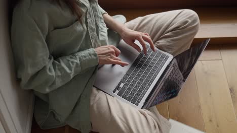 woman working on a laptop sitting on a wooden floor near a window