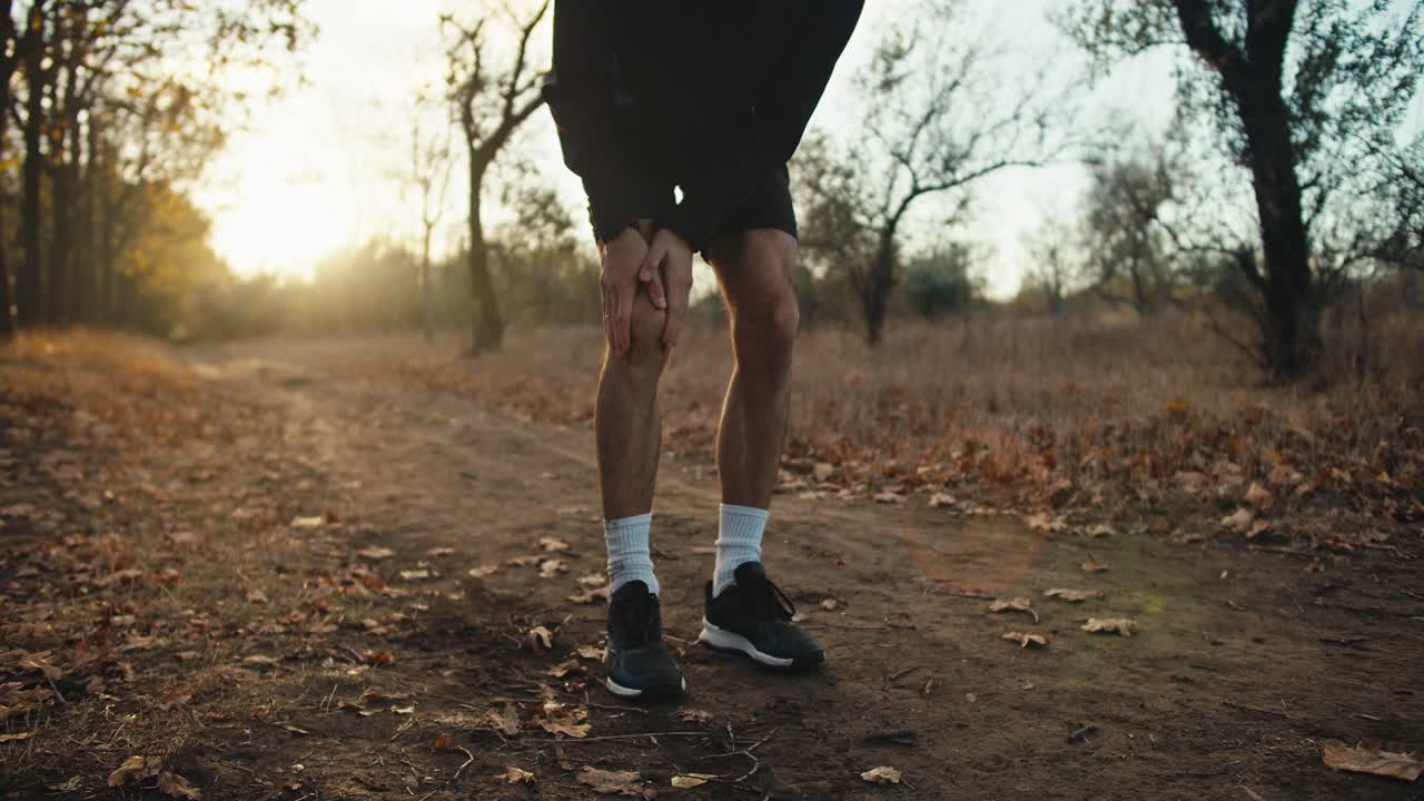 A guy an athlete in a black sports uniform while jogging in the forest holds his knee and rubs it because he feels damage and injury