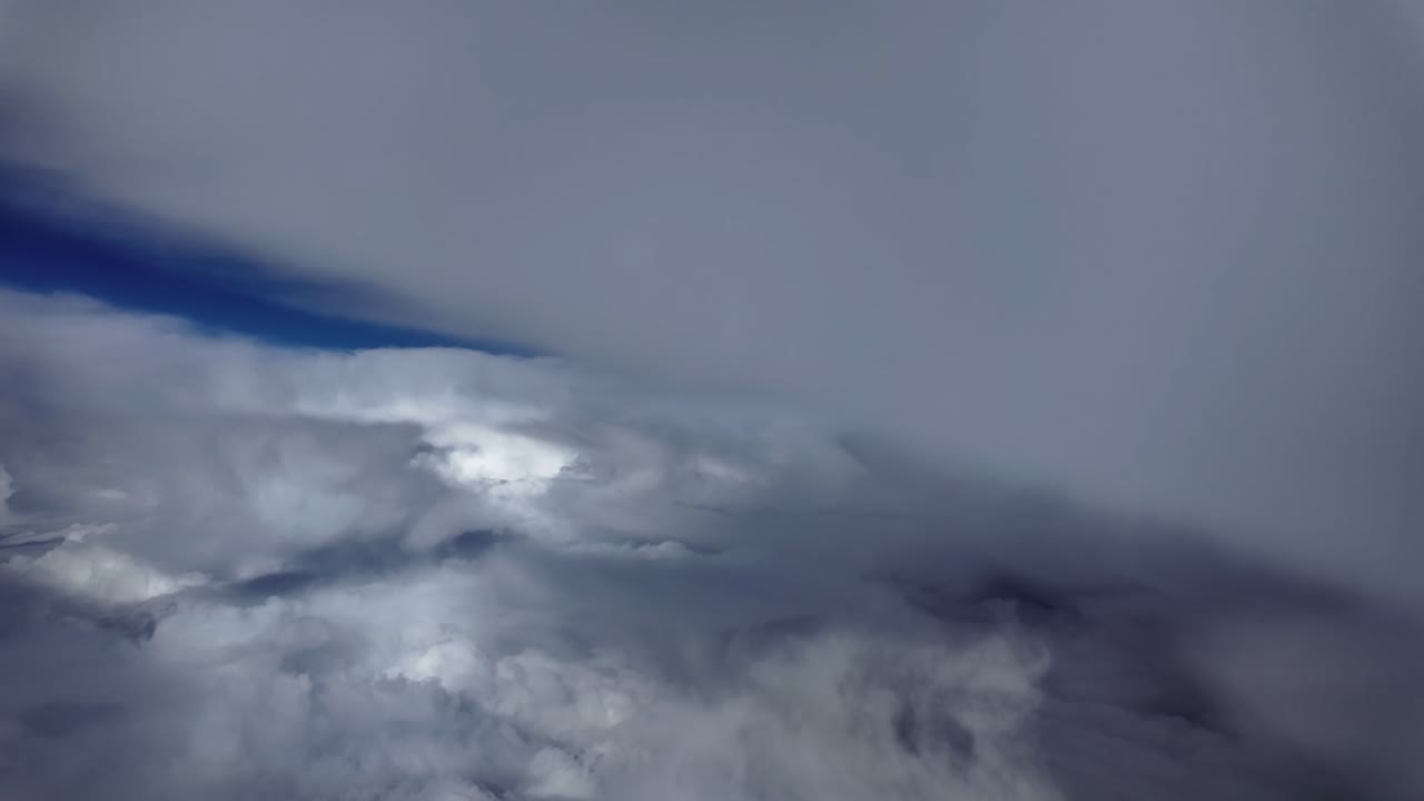 Et pilotperspektiv filmet fra cockpittet i et jetfly, der flyver i marchhøjde gennem en himmel fuld af truende skyer i en intens blå farve i baggrunden.