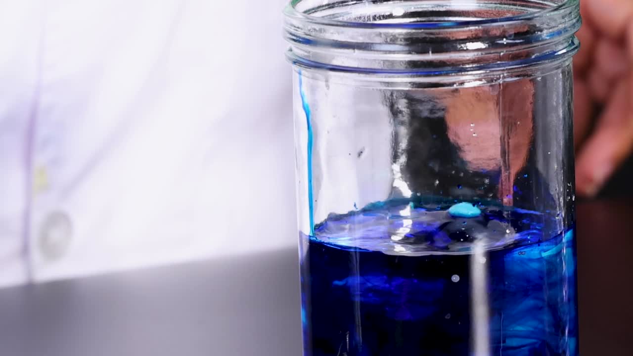 A blue liquid in a glass jar reacts, creating bubbles and a smoky effect.