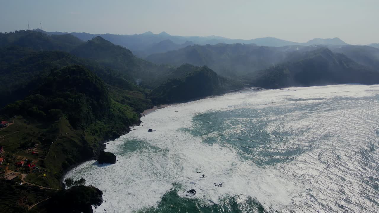 Cinematic drone shot of giant waves of Ocean reaching beautiful coastline with mountain range in summer - Central Java, Indonesia - Establishing drone flight