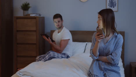 hombre y mujer guapos recién casados en pijama desplazándose y escribiendo en sus teléfonos inteligentes mientras se sientan en las diferentes esquinas de la cama por la noche